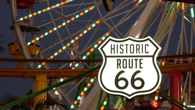 SANTA MONICA, LOS ANGELES, USA - 28 OCT 2019: Iconic road sign glowing, historic route 66. Famous california symbol, pier of pacific ocean resort. Illuminated festive ferris wheel in amusement park.