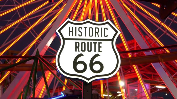 SANTA MONICA, LOS ANGELES, USA - 28 OCT 2019: Iconic road sign glowing, historic route 66. Famous california symbol, pier of pacific ocean resort. Illuminated festive ferris wheel in amusement park.