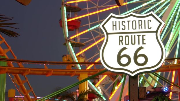 SANTA MONICA, LOS ANGELES, USA - 28 OCT 2019: Iconic road sign glowing, historic route 66. Famous california symbol, pier of pacific ocean resort. Illuminated festive ferris wheel in amusement park.