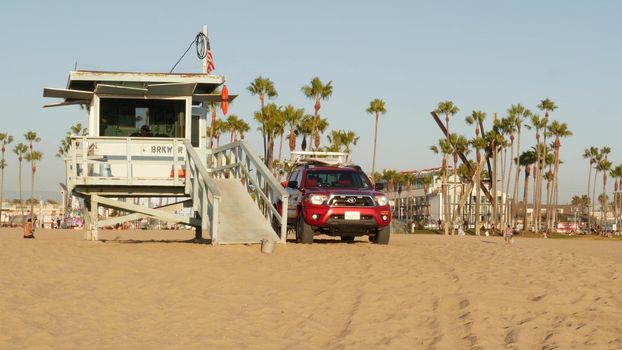 LOS ANGELES CA USA - 16 NOV 2019: California summertime Venice beach aesthetic. Iconic retro wooden lifeguard watchtower, baywatch red car. Life buoy, american state flag and palms near Santa Monica.