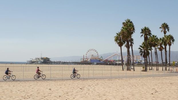 SANTA MONICA, LOS ANGELES CA USA - 28 OCT 2019: California summertime beach aesthetic, people walking and ride cycles on bicycle path. Amusement park on pier and palms. American pacific ocean resort.