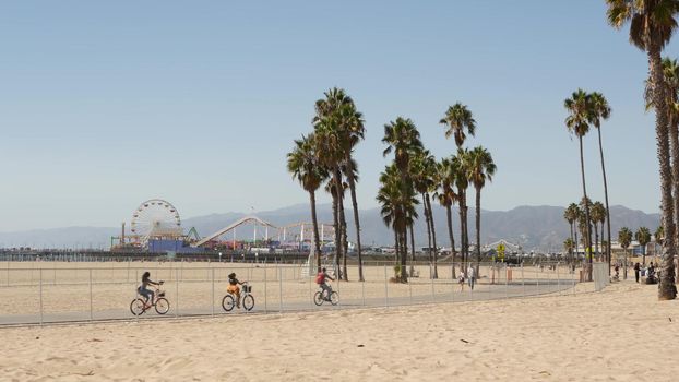 SANTA MONICA, LOS ANGELES CA USA - 28 OCT 2019: California summertime beach aesthetic, people walking and ride cycles on bicycle path. Amusement park on pier and palms. American pacific ocean resort.