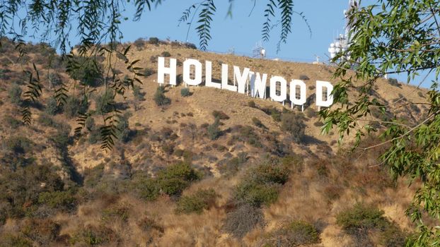 LOS ANGELES, CALIFORNIA, USA - 7 NOV 2019: Iconic Hollywood sign. Big letters on hills as symbol of cinema, movie studios and entertainment industry. Large text on mountain, view thru green leaves.