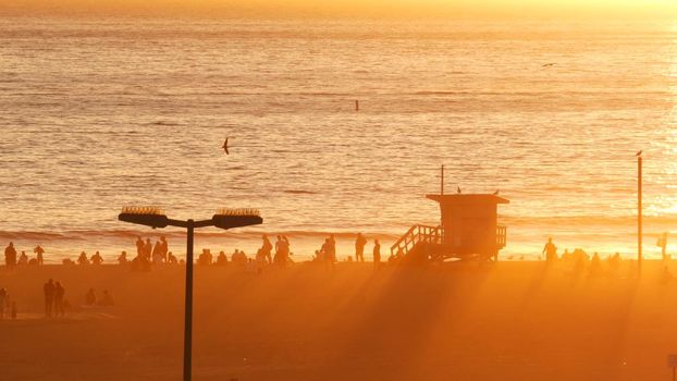 SANTA MONICA, LOS ANGELES, USA - 28 OCT 2019: California summertime beach aesthetic, atmospheric golden sunset. Unrecognizable people silhouettes, sun rays over pacific ocean waves. Lifeguard tower.