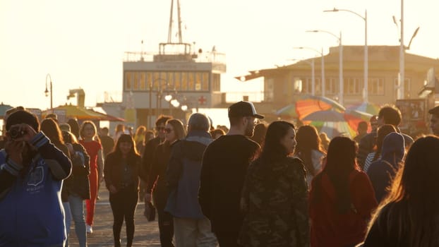 SANTA MONICA, LOS ANGELES CA USA - 19 DEC 2019: Many multiracial people walking on pier. Pedestrians walk, overcrowded seafront promenade. Crowd in golden sun light on broadwalk, sun rays over heads.