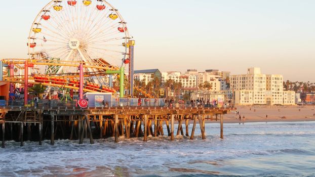 SANTA MONICA, LOS ANGELES CA USA - 19 DEC 2019: Classic ferris wheel in amusement park on pier. California summertime beach aesthetic, ocean waves in pink sunset. Summertime iconic symbol.