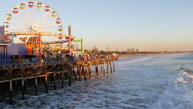 SANTA MONICA, LOS ANGELES CA USA - 19 DEC 2019: Classic ferris wheel in amusement park on pier. California summertime beach aesthetic, ocean waves in pink sunset. Summertime iconic symbol.