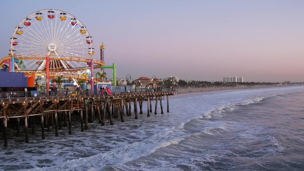 SANTA MONICA, LOS ANGELES CA USA - 19 DEC 2019: Classic illuminated ferris wheel in amusement park on pier. California summertime beach aesthetic, ocean waves in pink sunset. Summertime iconic symbol.