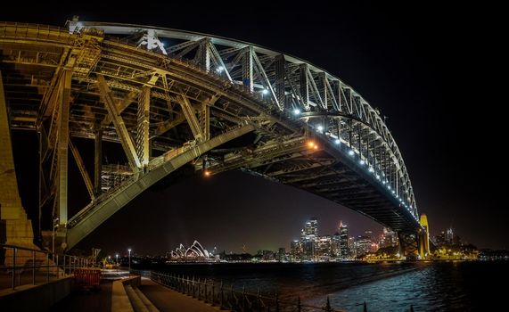 Ostseite der Hafenbrücke von Sydney bei Sonnenuntergang mit heller Beleuchtung des Stahlbogens und der Säulen, die im verschwommenen Wasser des Hafens mit CBD der Stadt Sydney im Hintergrund reflektieren.