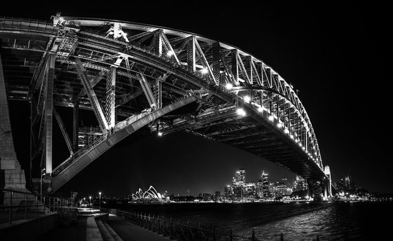 black and white East side of Sydney harbour bridge at sunset with bright illumination of steel arch and columns reflecting in the blurred waters of harbour with Sydney city CBD in the background.