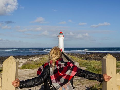 A young woman dressed in black is looking towards a white lighthouse while holding on a fence with her hands, Great Ocean Road, Australia