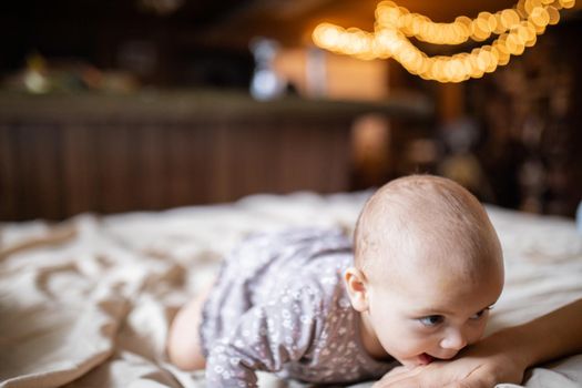 Adorable and happy baby lying on her stomach above bed with blurry background. Lovely baby on bed gently biting the hand of her mother. Cute toddlers