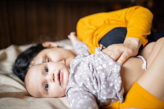 Cute and happy baby lying down on bed next to her sleeping mother. Young woman wearing yellow clothing on bed and tenderly hugging adorable baby. Lovely maternity