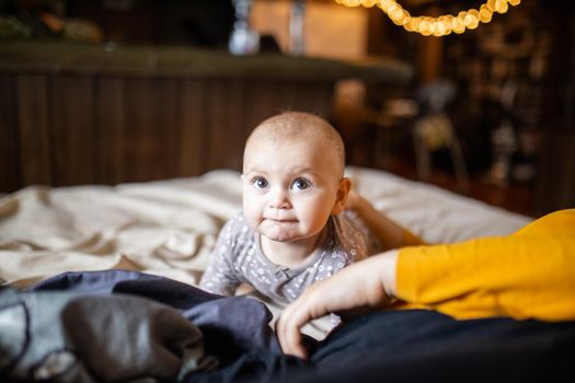 Adorable and curious-looking baby lying on her stomach above bed with blurry background. Lovely baby on bed with cream color sheets next to her mother. Cute toddlers