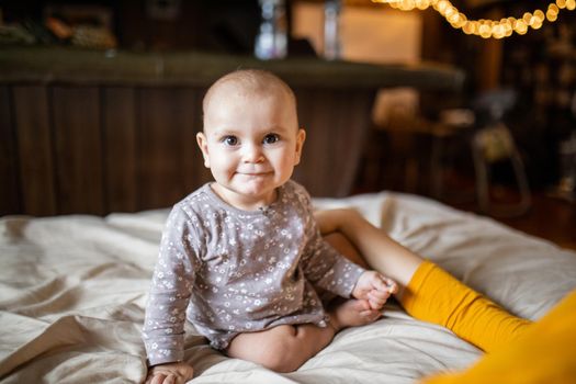 Adorable and happy baby sitting on bed with blurry background. Lovely baby on bed with cream color sheets next to her mother. Cute toddlers