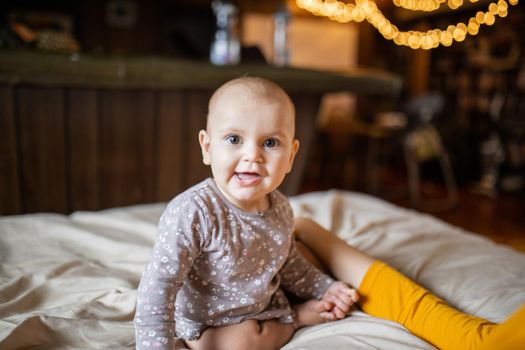 Adorable and happy baby sitting on bed with blurry background. Lovely baby on bed with cream color sheets next to her mother. Cute toddlers