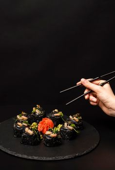 Hand with chopsticks wants to take custom sushi roll with black rice, crab meat, avocado, smoked salmon mousse, oar caviar, masago, shrimp cocktail, edible gold leaf, ginger, wasabi on black table