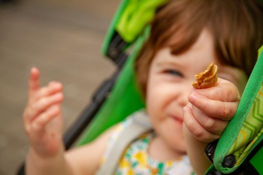 Portrait of a cute, blue-eyed, brown-haired baby girl sitting in a green pushchair holding a piece of waffle towards the camera while eating