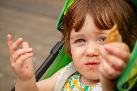 Portrait of a cute, blue-eyed, brown-haired baby girl sitting in a green pushchair holding a piece of waffle towards the camera while eating