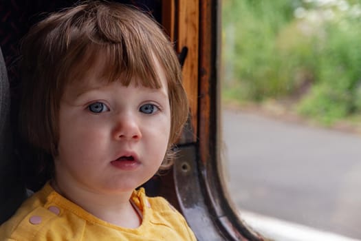Portrait of a cute, blue-eyed, brown-haired baby girl in a yellow shirt sitting on a window