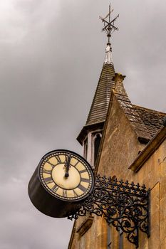 An old iron clock hanging on a church stone wall with a steeple in the background on a cloudy, grey day.