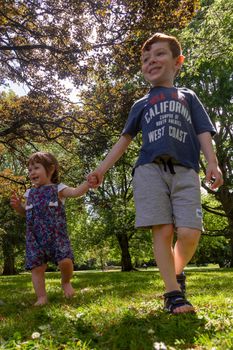 A cute, blue-eyed, red-haired, 4 years old boy and a cute, blue-eyed, brown-haired baby girl walking in a park holding hands on a sunny summer day wearing shorts.