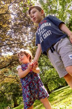 A cute, red-haired, blue-eyed, 4 years old child and a cute, blue-eyed, brown-haired baby girl standing in a park holding hands on a sunny day