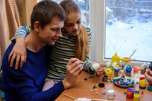 Father and daughter paint easter eggs while sitting at the table by the window