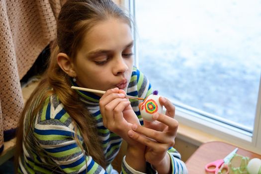 Girl paints Easter eggs while sitting at the table by the window