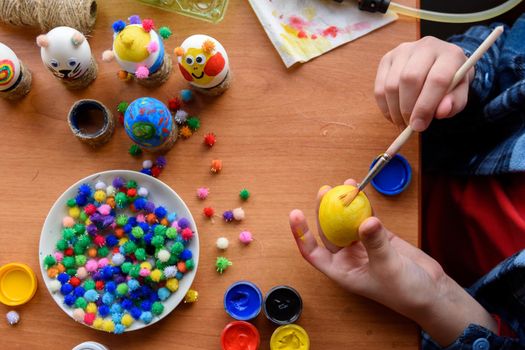 Top view of the desk and the hands of a child who paints festive easter eggs with brushes