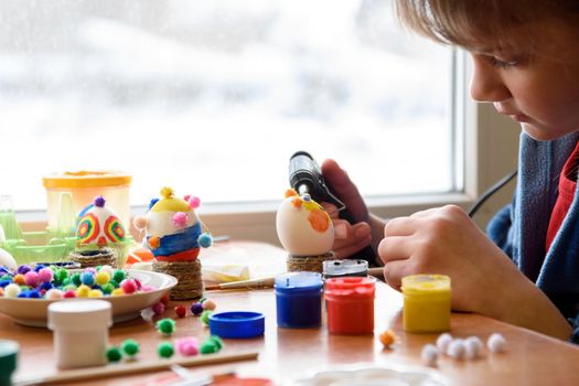 A girl glues elements to an Easter egg with a glue gun