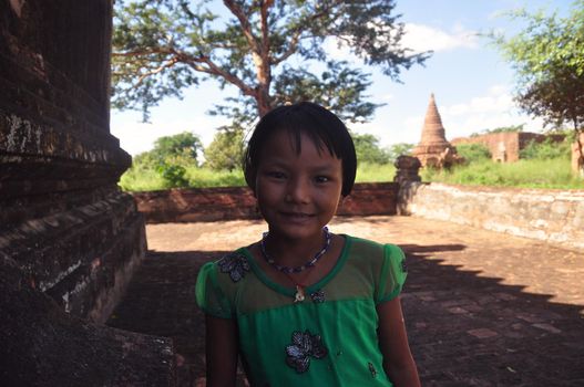 BAGAN, MYANMAR - NOVEMBER 18, 2015 Portrait of adorable little ethnic girl in stone yard of tropical city looking at camera, Myanmar. Charming little ethnic girl outdoors