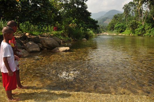 NAKHONSITHAMMARAT, THAILAND - JUNE 7, 2014: Kids in shallow clear water of pond, View of little ethnic boys standing in clear shallow water of flow with fish splashing. Mountain on background