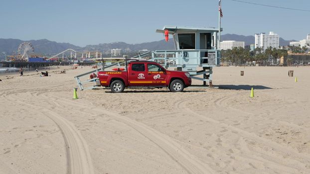 SANTA MONICA, LOS ANGELES CA USA - 28 OCT 2019: California summertime beach aesthetic. Iconic blue wooden watchtower, red lifeguard car on sandy sunny coast. Amusement park and attractions on the pier