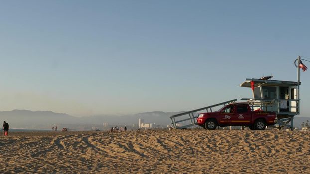 LOS ANGELES CA USA - 16 NOV 2019: California summertime Venice beach aesthetic. Iconic retro wooden lifeguard watchtower, baywatch red car. Life buoy and american state flag near Santa Monica.