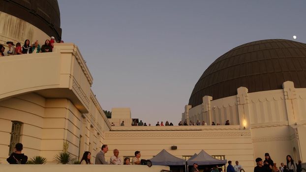 LOS ANGELES, CALIFORNIA, USA - 7 NOV 2019: Griffith observatory viewpoint. Crowd on vista point, people watching sunset over city and Hollywood sign. Many multiracial tourists look at golden sundown.