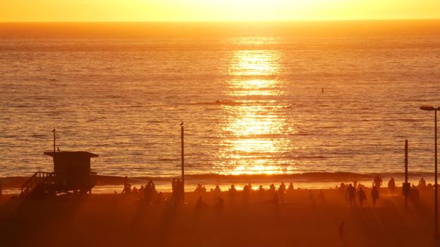 SANTA MONICA, LOS ANGELES, USA - 28 OCT 2019: California summertime beach aesthetic, atmospheric golden sunset. Unrecognizable people silhouettes, sun rays over pacific ocean waves. Lifeguard tower.