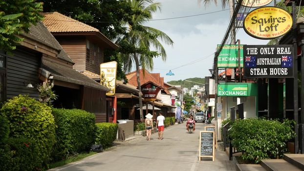 SAMUI ISLAND, THAILAND - MAY 27, 2019: Typical touristic street in Fisherman village with souvenir stores. View of calm lane of city in Asia with touristic shops in daytime. Thai people on motorbikes