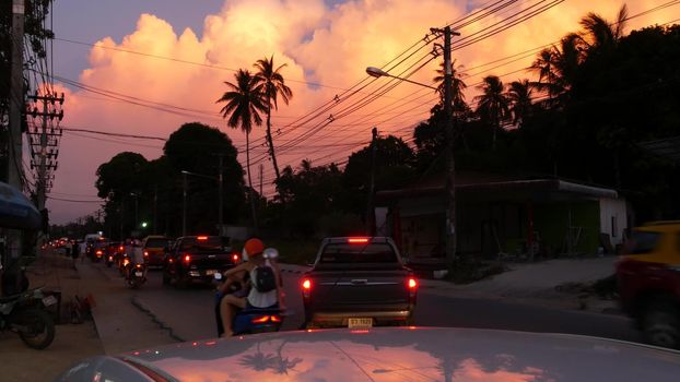 KOH SAMUI ISLAND, THAILAND - 10 JULY 2019: City road with urban transport moving. Back view of cars and motorbikes moving on busy road under orange clouds in sunset tropical exotic evening.