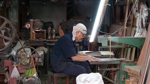 BANGKOK, THAILAND - 11 JULY, 2019: Ethnic craftsman working in street workshop. Side view of male artisan sitting near workbench and working in grungy workshop on street.