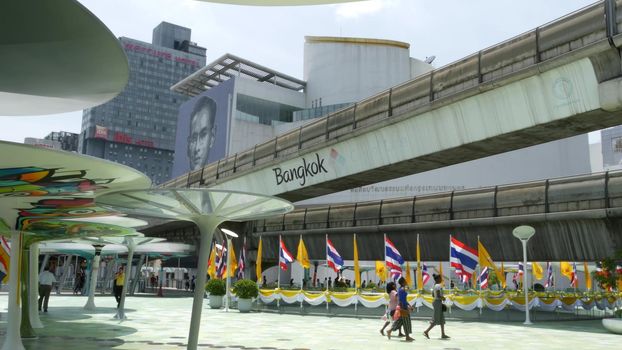 BANGKOK, THAILAND - 11 JULY, 2019: Pedestrians walking on the bridge near MBK and Siam Square under BTS train line. People in festive modern city decorated with waving national flags and royal symbol.