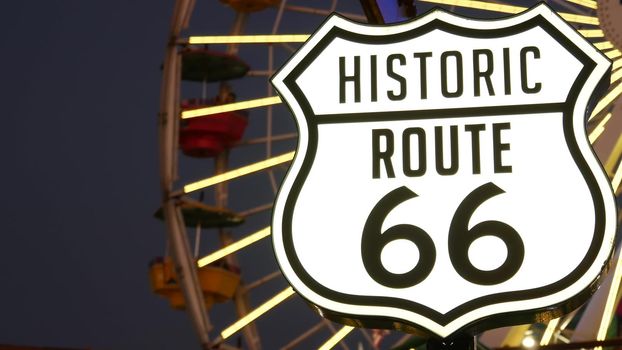 SANTA MONICA, LOS ANGELES, USA - 28 OCT 2019: Iconic road sign glowing, historic route 66. Famous california symbol, pier of pacific ocean resort. Illuminated festive ferris wheel in amusement park.