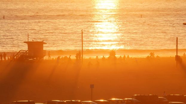 SANTA MONICA, LOS ANGELES, USA - 28 OCT 2019: California summertime beach aesthetic, atmospheric golden sunset. Unrecognizable people silhouettes, sun rays over pacific ocean waves. Lifeguard tower.