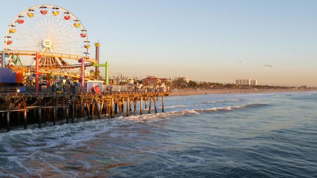 SANTA MONICA, LOS ANGELES CA USA - 19 DEC 2019: Classic ferris wheel in amusement park on pier. California summertime beach aesthetic, ocean waves in pink sunset. Summertime iconic symbol.