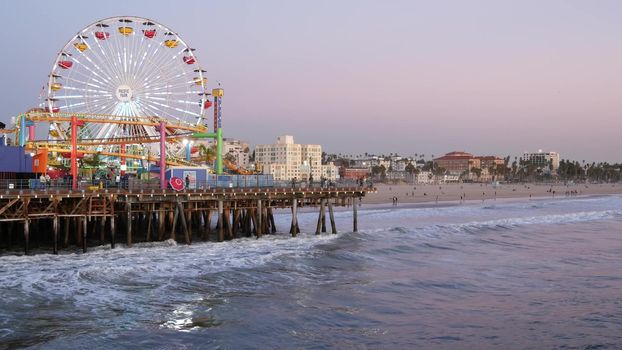 SANTA MONICA, LOS ANGELES CA USA - 19 DEC 2019: Classic illuminated ferris wheel in amusement park on pier. California summertime beach aesthetic, ocean waves in pink sunset. Summertime iconic symbol.