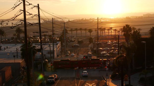 SAN DIEGO, CALIFORNIA USA - 15 JAN 2020: Level crossing near Lindbergh field international airport. Railroad red tram and runway at sunset. MTS Trolley rail transportation, cars and railway transport.