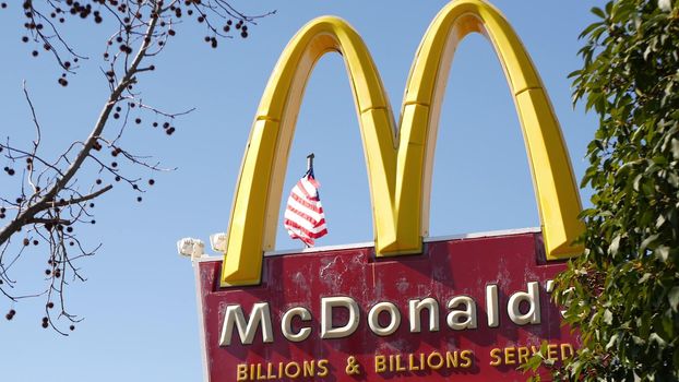 VISTA, CALIFORNIA USA - 16 FEB 2020: McDonalds logo sign and American flag. Yellow letter M icon and Stars and Stripes. Golden Arches emblem, brand sign in suburban district. Food restaurant on street
