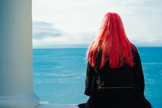 A girl with pink hair looks out at the sea by a white column.