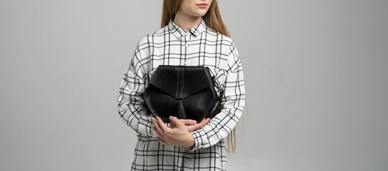 Small black leather bag in a woman's hand on a white background. Shoulder handbag. Woman in a white plaid shirt and black jeans and with a black handbag. Style, retro, fashion, vintage and elegance