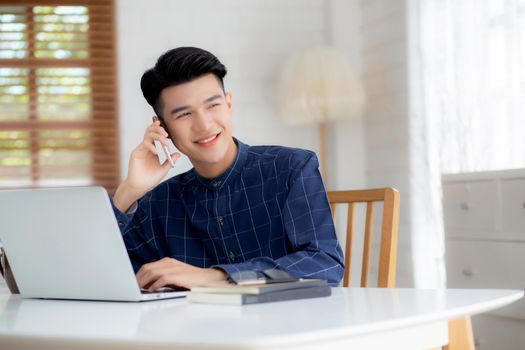 Young asian man talking phone and work from home with laptop computer, freelance using notebook to internet online and speak on smartphone, domestic life, business and communication concept.